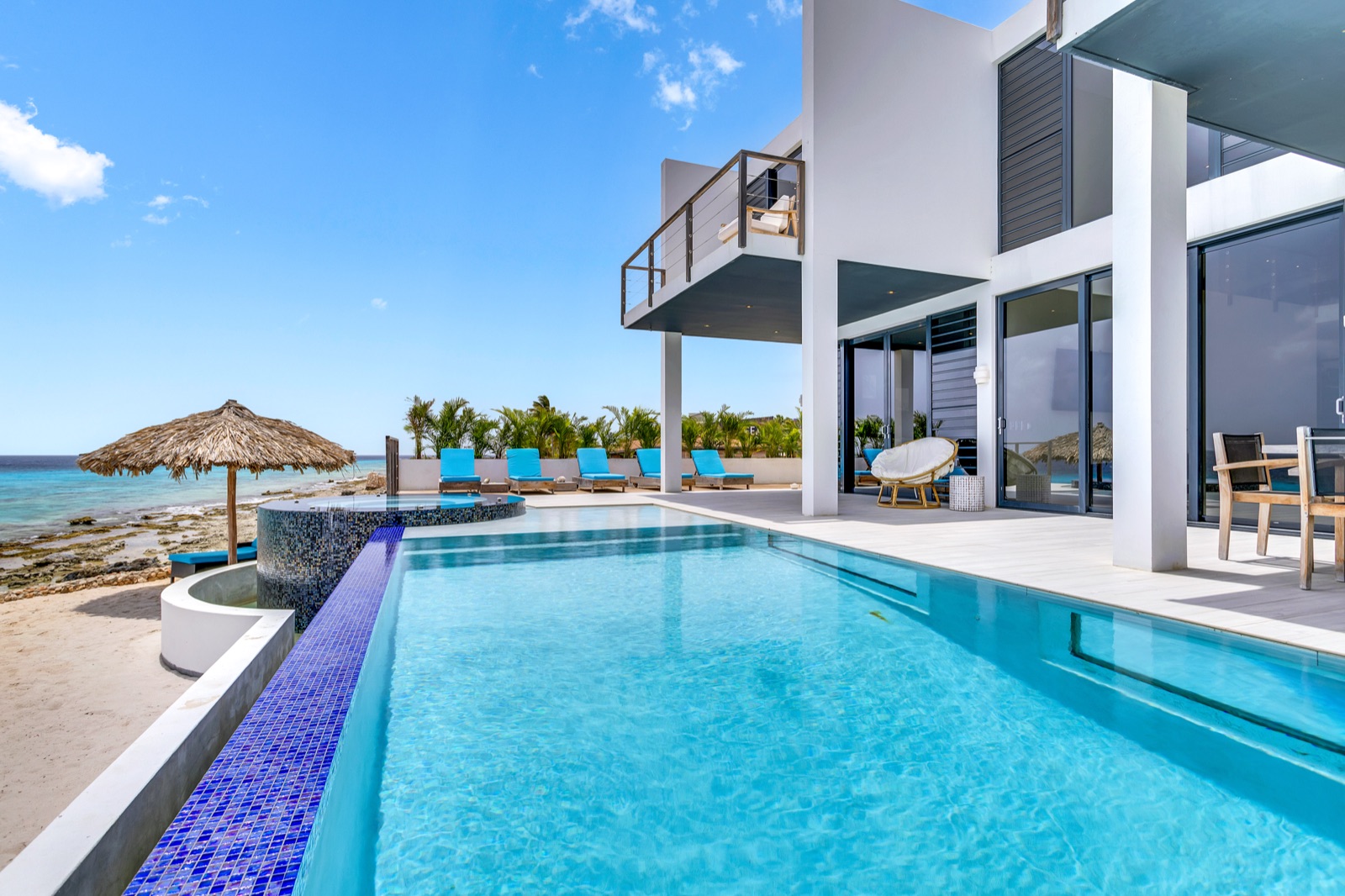 Luxury Bonaire villa with a modern white exterior, a private infinity pool with blue mosaic tiles, and direct access to a sandy and rocky beach on the Caribbean Sea.